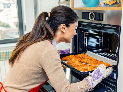 Woman leaning towards the open oven and taking a baking pan with croissants out of it
