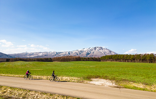A couple are riding their bikes together in the countryside while enjoying views of a vast grassy field and Mt. Iwate off in the distance.
