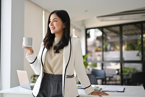 Attractive Asian businesswoman in her office daydreaming while sipping coffee.