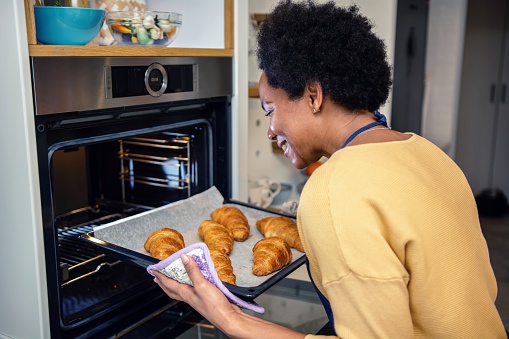 Black woman leaning towards the open oven and taking a baking pan with croissants out of it