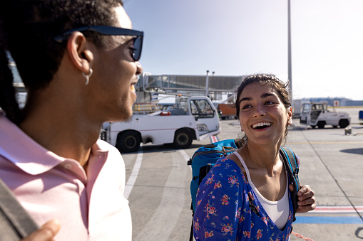 A side-view shot of a young man and woman walking in a queue outdoors on an airport runway in Toulouse, France waiting to get on their flight. They are looking at each other and smiling.
