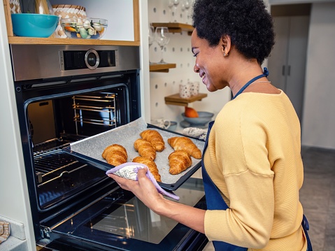 Black woman leaning towards the open oven and taking a baking pan with croissants out of it