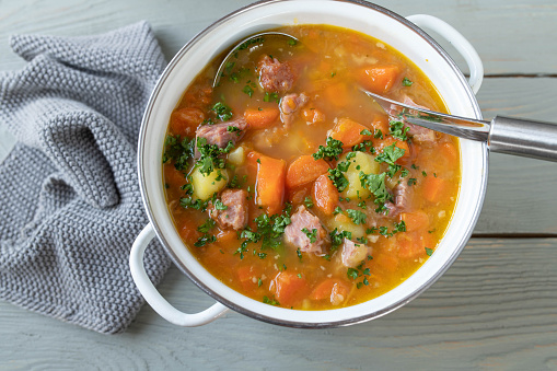 Delicious vegetables stew with carrots, potatoes and smoked pork meat. Served in white enamel pot with ladle and garnish isolated on light wooden table background from above.