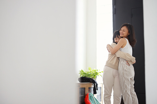 Standing in the hallway in front of the door, an Asian mom affectionately hugged her daughter before she got ready to go to school, in a display of love, care, and family bonding.