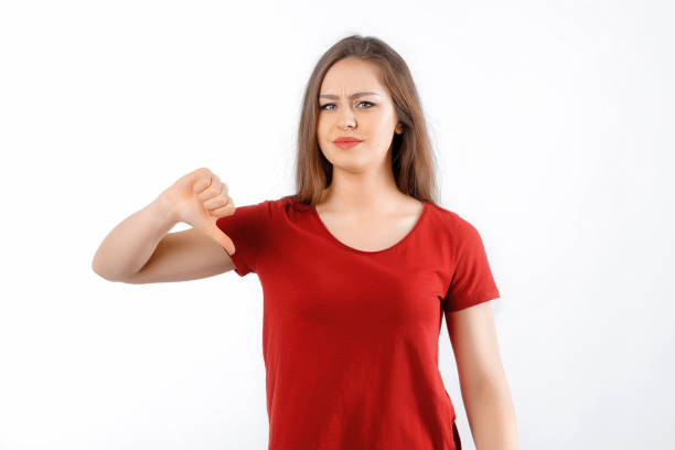 istock Discontent young woman shows disapproval sign, keeps thumb down, expresses dislike, frowns face in discontent, dressed in red t shirt, standing over white background. Body language concept 1477437053