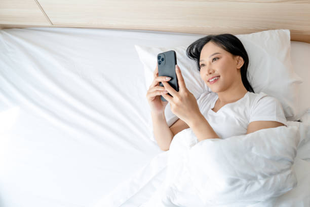 istock Beautiful Asian woman using smartphone on white bed in morning with copy space. Mobile addiction concept. 1477443800