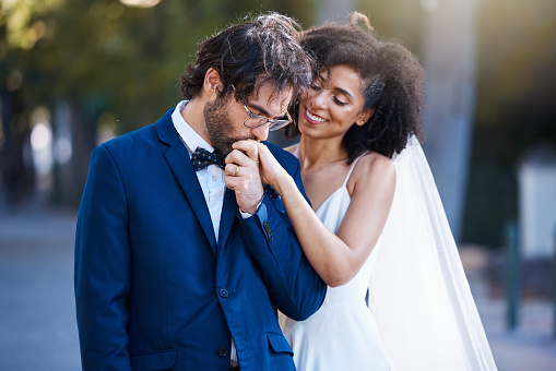 Wedding, happy couple and kiss on hand at marriage celebration event together with commitment. Interracial man and woman at ceremony with trust, partnership and care while outdoor with flower bouquet