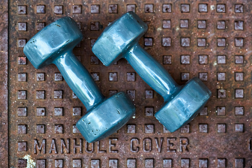 Medium-weight dumbbells, worn and well-used, resting outdoors on a highly textured metal cover made of cast iron.