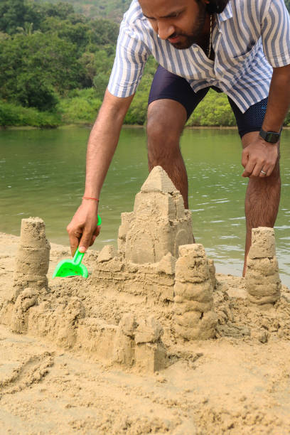 Stock photo showing a man bent down building a complex sandcastle built on the beach in Goa, India. This structure was made with a bucket and green spade, with an exterior wall modification to turn it into a fortress