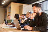 Young businessman yawning while working on a laptop in a busy office lounge