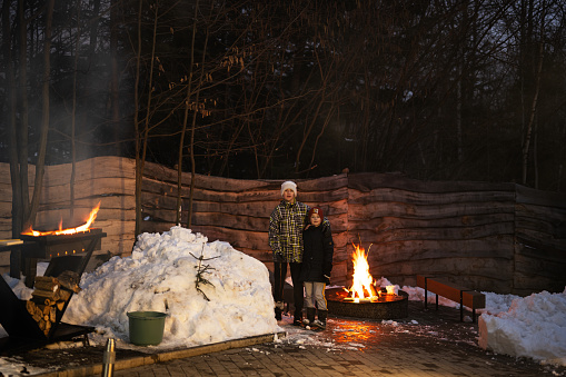 Two brothers stand together by the fire pit in winter night.