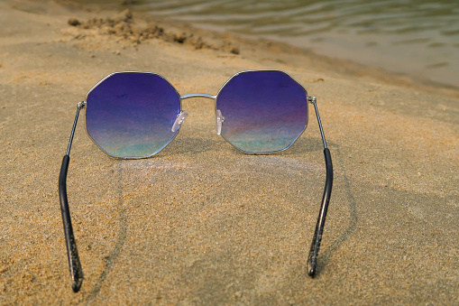 Stock photo showing close-up view of sandy beach with a pair of octagonal, metal framed, tinted, mirrored sunglasses on damp sand at sea water's edge at low tide, against a backdrop of woodland with palm trees.