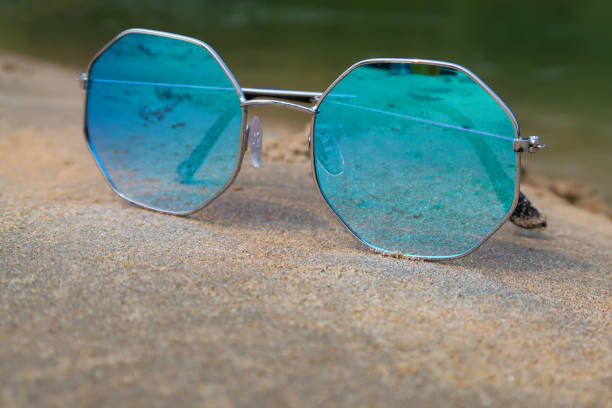istock Close-up image of pair octagonal, metal framed, tinted, mirrored sunglasses on damp beach sand, sea background at low tide, focus on foreground 1477463052