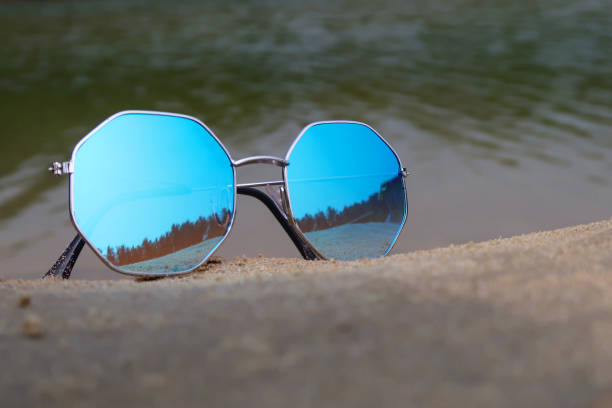istock Close-up image of pair octagonal, metal framed, tinted, mirrored sunglasses on damp beach sand, sea background at low tide, focus on foreground 1477463053