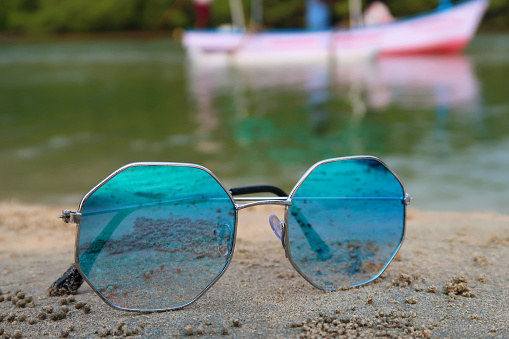 Stock photo showing close-up view of sandy beach with a pair of octagonal, metal framed, tinted, mirrored sunglasses on damp sand at sea water's edge at low tide, against a backdrop of woodland with palm trees.