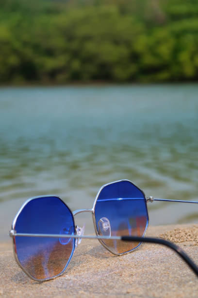 istock Close-up image of pair octagonal, metal framed, tinted, mirrored sunglasses with unfolded arms on damp beach sand, coastal background of sea and woodland with palm trees, rear view, focus on foreground, copy space 1477463066