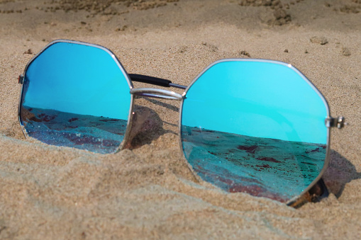 Stock photo showing close-up view of sandy beach with a pair of octagonal, metal framed, tinted, mirrored sunglasses on sandy beach.