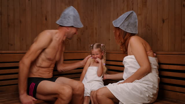 Loving father and mother kissing cheeks of smiling daughter sitting on wooden bench in Finn sauna. Happy carefree relaxed Caucasian family enjoying leisure in SPA indoors.