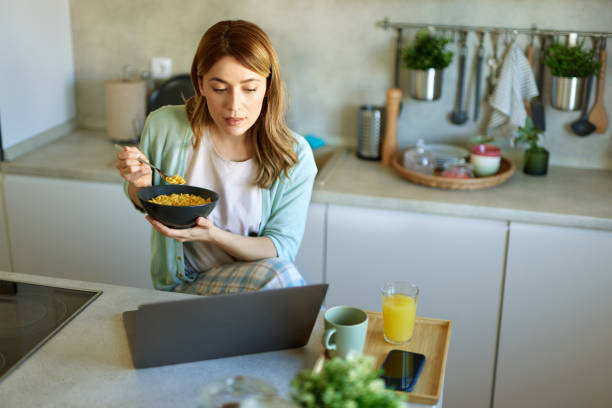 istock Young cheerful woman using laptop computer and eating cornflakes in the kitchen 1477471382
