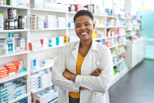 Mid adult pharmacist standing with arms crossed in front of shelves in pharmacy and smiling at camera.