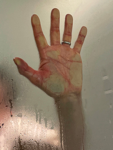 Stock photo showing close-up view of unrecognisable man behind a steamed-up shower cubicle glass screen. His hand is pictured reaching out with the fingers spread and the palm pushing against the glass making the scene appear mysterious and a little frightening.