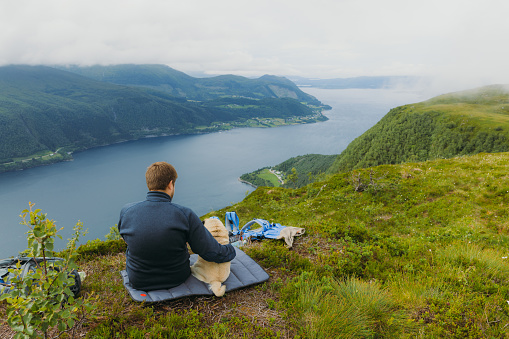Smiling Man backpacker with his cute dog relaxing on top of the mountain looking at dramatic summer landscape of Norway's nature in Scandinavia