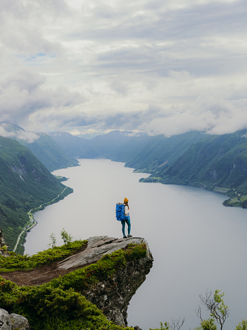 Happy female hiker in yellow hat getting to the edge of the cliff contemplating dramatic view of sea and green hills of the beautiful fjord in Western Norway