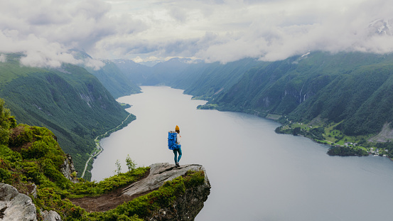 Happy female hiker in yellow hat getting to the edge of the cliff contemplating dramatic view of sea and green hills of the beautiful fjord in Western Norway