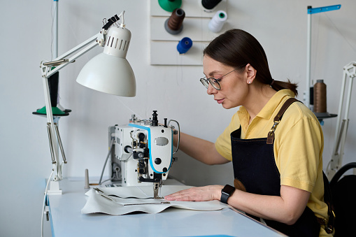Side view of brunette female tailor or fashion designer bending over electric sewing machine while sitting by workplace in atelier