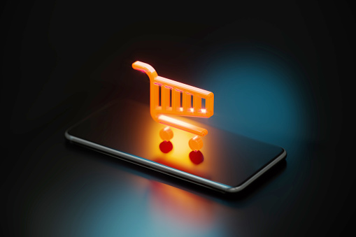 Yellow shopping cart symbol glowing over a smart phone on black background. Horizontal composition with copy space.