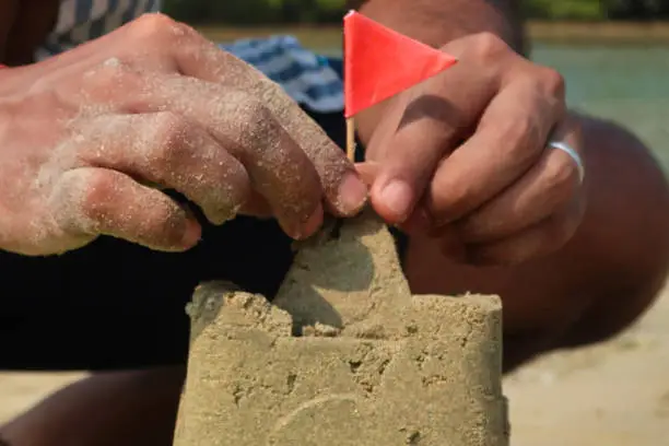 Stock photo showing sandy beach with a sandcastle made with a bucket with a mold shape, a pair of hands are seen making this sandcastle, fixing a red flag on a cocktail stick