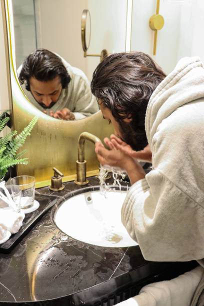 istock Image of Indian man stood bent at bathroom sink wearing bathrobe using hands to splash water on face, reflection in mirror, artificial fern plant in metal pot, toothbrushes in glass, self care and grooming concept 1477496497