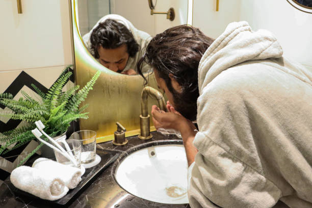 istock Close-up image of Indian man stood bent at bathroom sink wearing bathrobe using hands to splash water on face, reflection in mirror, artificial fern plant in metal pot, toothbrushes in glass, self care and grooming concept 1477496503