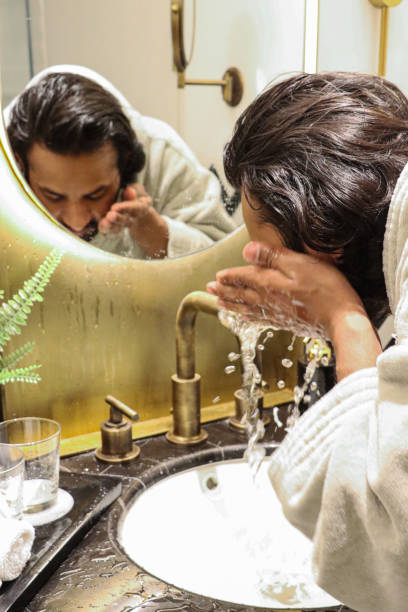 istock Image of Indian man stood bent at bathroom sink wearing bathrobe using hands to splash water on face, reflection in mirror, artificial fern plant in metal pot, toothbrushes in glass, self care and grooming concept 1477496505
