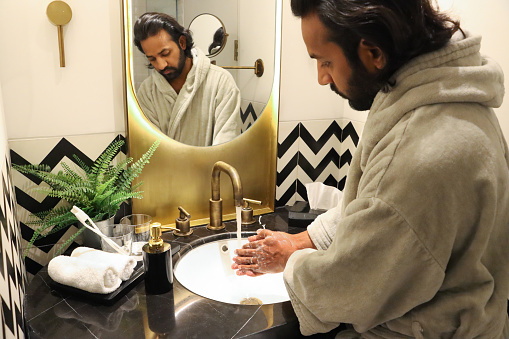 Stock photo showing luxury bathroom, with black and white zig-zag patterned wall tiles, white ceramic sink with dual knob monobloc mixer tap in front of mirror reflecting an Indian man wearing grey bathrobe whilst washing his hands under a flowing water tap. Self care and grooming concept.