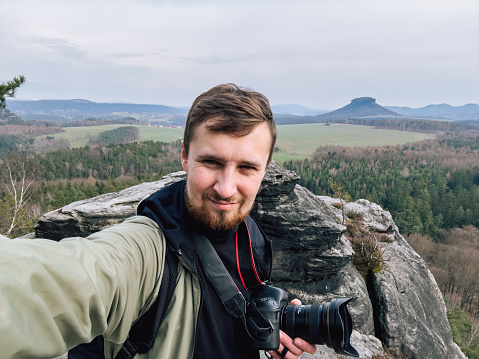 Male portrait of caucasian man outdoors who enjoys by view in the mountains. Lifestyle photography of real people. Person have backpack and relaxing by nature. Photographer is doing selfie in beautiful landscape