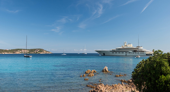 This stunning wide-angle view captures the beautiful vessels on the water at the luxurious Sardinia resort in Sardinia, Italy. The expansive view showcases a massive super-yacht on the right, contrasted by the smaller vessels nearby, highlighting the sheer size and grandeur of the larger boat. The breathtaking scenery of the Mediterranean is visible in the background, with the sun-drenched hills and lush vegetation framing the tranquil turquoise and blue waters of the bay. This image perfectly captures the summertime paradise and exclusive luxury destination that Sardinia is known for. Visitors to this awesome location can immerse themselves in the beauty of the Mediterranean, enjoy luxurious accommodations, and experience the ultimate in relaxation and serenity.
