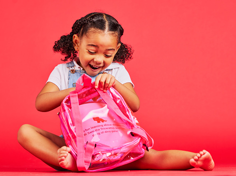 School, bag and red background with a student black girl in studio sitting on the floor against a wall. Children, education and excited with a female kid pupil getting ready for learning, or growth