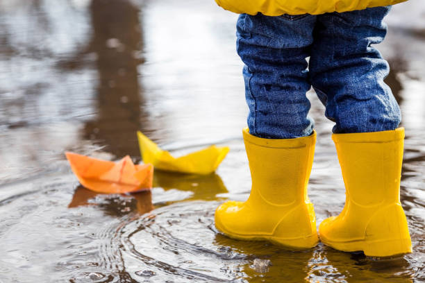 es ist frühling. ein kleines kind in gelben gummistiefeln springt durch pfützen, spielt und lässt papierboote auf dem wasser zu wasser. foto von frühlings- und herbstferien. das konzept des frühlings. - puddle rain boot water stock-fotos und bilder