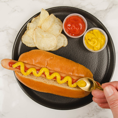 A hot dog, sausage in a sliced bread bun, with ketchup and mustard, on a black plate. A hand with a golden spoon putting mustard on the sausage. Potato chips and two small bowls with red ketchup and yellow mustard are also on the plate. Plate standing on a white marble table. Top view.