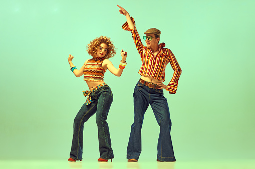 Two excited people, man and woman in retro style clothes dancing disco dance over green background. 1970s, 1980s fashion, music, hippie lifestyle,