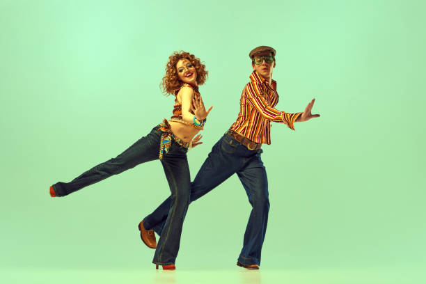 복고풍 컬러 셔츠와 플레어 청바지를 입은 행복한 남자와 여자의 역동적인 초상화는 녹색 배경 위에 활기찬 춤을 추고 있습니다. 70 년대, 1980 년대, 음악의 패션 트렌드의 개념 - 20 25 years 뉴스 사진 이미지