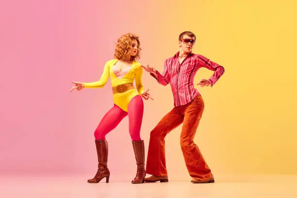Photo of Young stylish emotional man and woman, professional dancers in retro style clothes dancing disco dance over pink-yellow background. 1970s, 1980s fashion, music concept