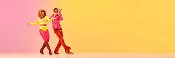 Photo of Stylish expressive excited couple of professional dancers in retro style clothes dancing disco dance over pink-yellow background. Concept of 70s, 80s fashion style, music and emotions