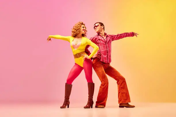 Photo of Disco dance. Stylish expressive excited couple of professional dancers in retro style clothes dancing over pink-yellow background.