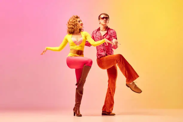 Photo of Disco style. Young emotional man and woman, professional dancers in retro clothes dancing dance over pink-yellow background. 1970s, 1980s fashion, music concept