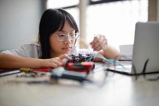 A young Asian girl is working on a robotic car for a science project. She is participating in her school's science and technology program through remote learning and using her laptop at home