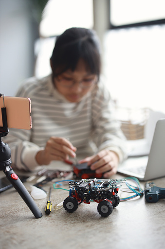 A young Asian girl is working on a robotic car for a science project. She is participating in her school's science and technology program through remote learning and using her laptop at home