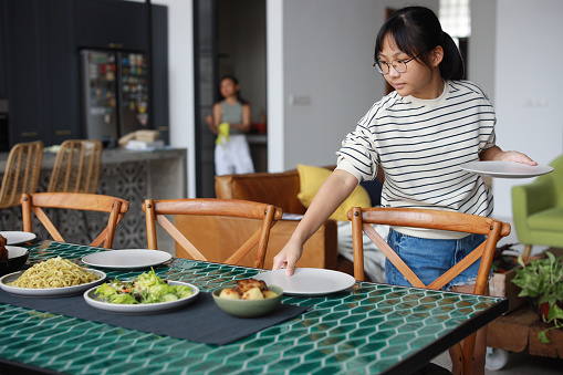 Young Asian girl putting plates on table together for lunch. A helping hand, teamwork and family concept