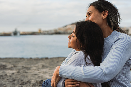 Happy latin mother hugging her little daughter on the beach while looking the sunrise together - Family and love concept